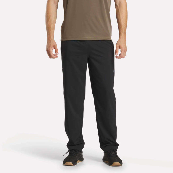 A person wearing a brown short-sleeve shirt, Reebok Men's Train Woven Unlined Pants, and black sneakers stands facing forward against a plain white background.