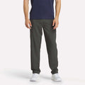 A man stands facing forward against a plain white background wearing Reebok Men's Train Woven Unlined Pants, a navy shirt, and white sneakers. The image shows him from shoulders to feet—ideal attire for gym workouts.
