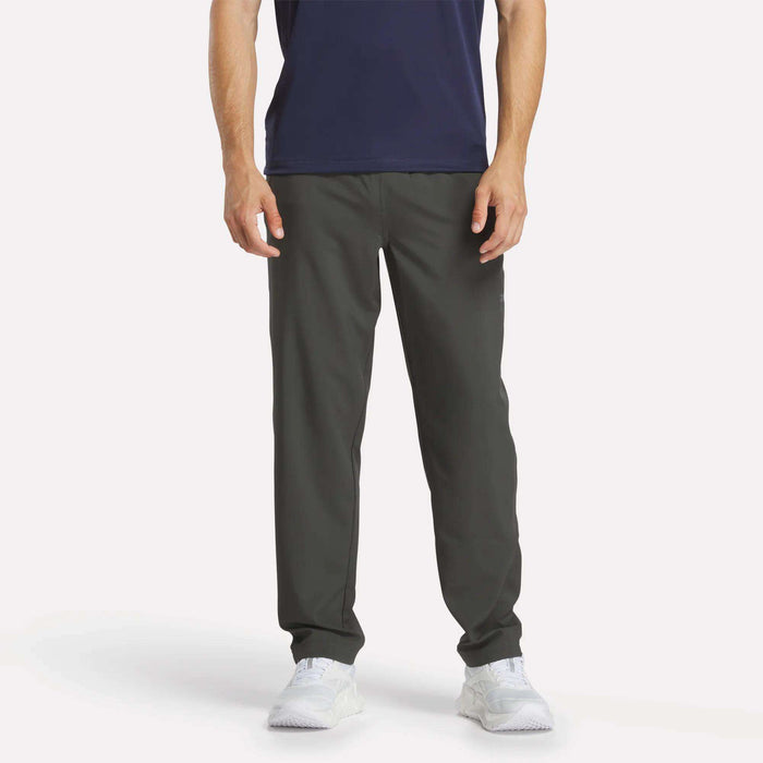 A man stands facing forward against a plain white background wearing Reebok Men's Train Woven Unlined Pants, a navy shirt, and white sneakers. The image shows him from shoulders to feet—ideal attire for gym workouts.