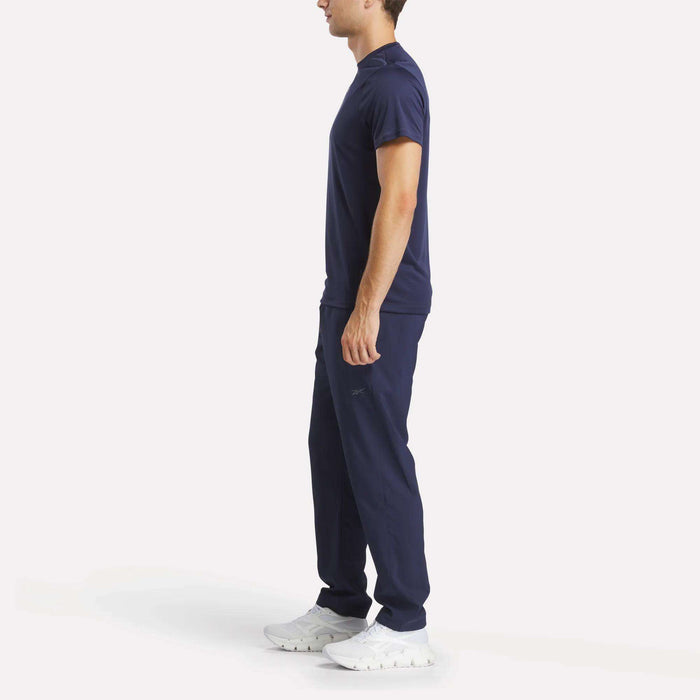 A man stands in profile against a white background, wearing Reebok Men's Train Woven Unlined Pants in navy blue with a matching t-shirt and white sneakers.