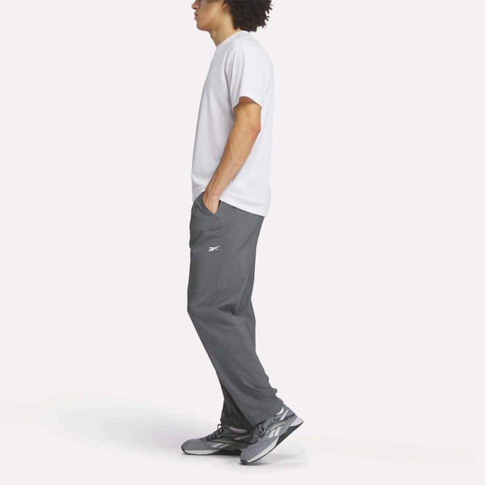 A person stands in profile against a plain white background, wearing a white t-shirt, gray Reebok Men's Train Woven Unlined Pants with a small logo, and gray sneakers.