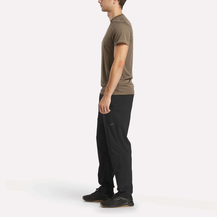 A man stands in profile facing left, wearing a brown t-shirt, black Reebok Men's Train Woven Unlined Pants, and black sneakers against a plain white background.