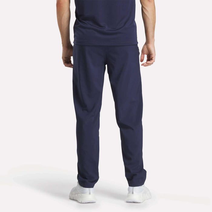 A person faces away, wearing Reebok Men's Train Woven Unlined Pants, a navy blue T-shirt, and white sneakers against a plain light background.