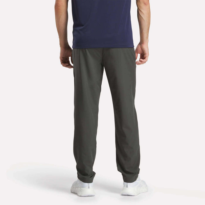 A person stands facing away, wearing a navy T-shirt, Reebok Men's Train Woven Unlined Pants ideal for gym sessions, and white sneakers against a plain white background.