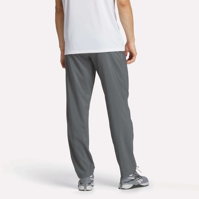 Someone stands facing away from the camera against a plain background, wearing a white t-shirt, gray sneakers, and Reebok Men's Train Woven Unlined Pants.