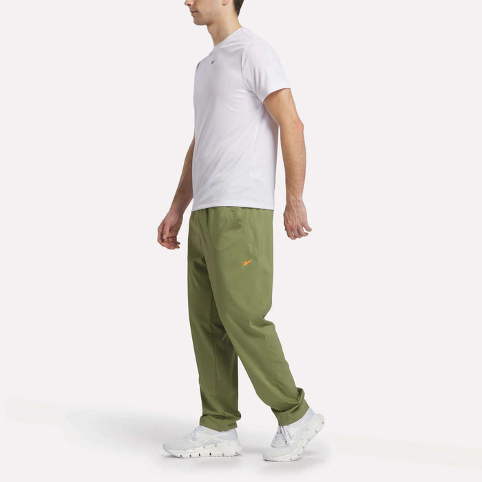 A man faces left against a plain white background, wearing a white t-shirt, white sneakers, and olive green Reebok Men's Train Woven Unlined Pants made from lightweight fabric with a small orange logo.