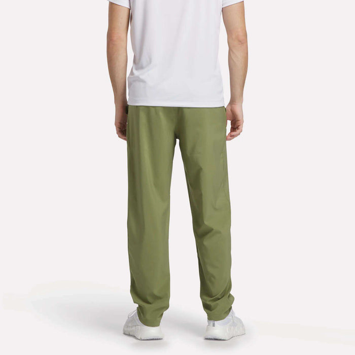 A person wearing a white short-sleeve shirt, Reebok Men's Train Woven Unlined Pants in light green, and white sneakers stands facing away from the camera against a plain white background.