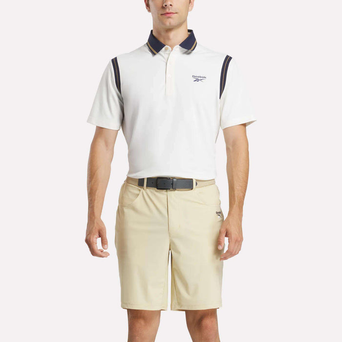 A man stands facing forward with hands relaxed, wearing a white Reebok Men's Varsity Polo Shirt with dark accents and beige shorts against a plain light background.
