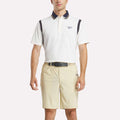A man stands facing forward with hands relaxed, wearing a white Reebok Men's Varsity Polo Shirt with dark accents and beige shorts against a plain light background.