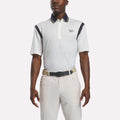 A man wearing a chalk-colored Men's Varsity Polo Shirt by Reebok, crafted from moisture-wicking RBK-DRY fabric and featuring navy accents, along with beige pants and a black belt, stands with his arms relaxed at his sides against a plain background.