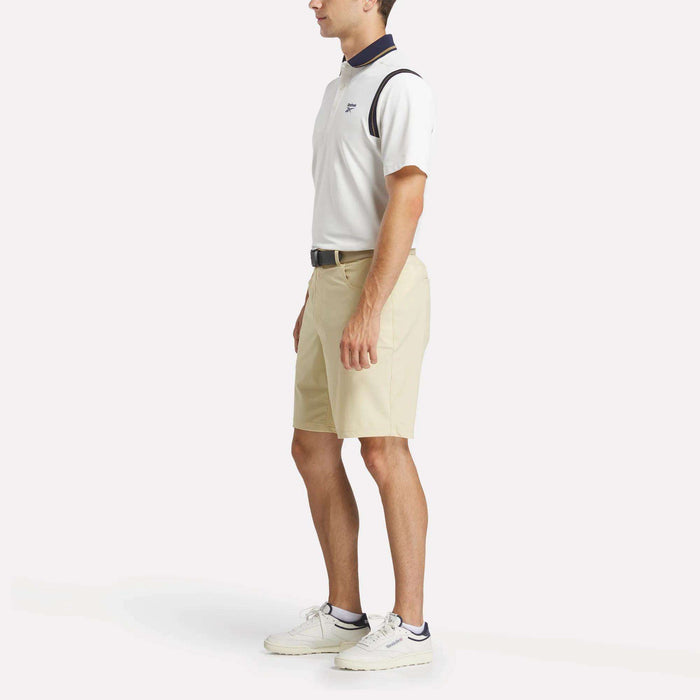 A man stands in side profile, wearing a men's Varsity Polo Shirt by Reebok, beige shorts, a black belt, white sneakers, and white ankle socks against a plain white background.