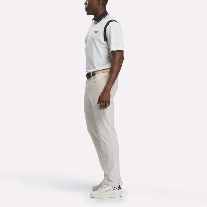 A man stands in profile wearing a crisp Reebok Men's Varsity Polo Shirt, paired with light beige pants and white sneakers. His hands are relaxed by his sides against a plain white background.