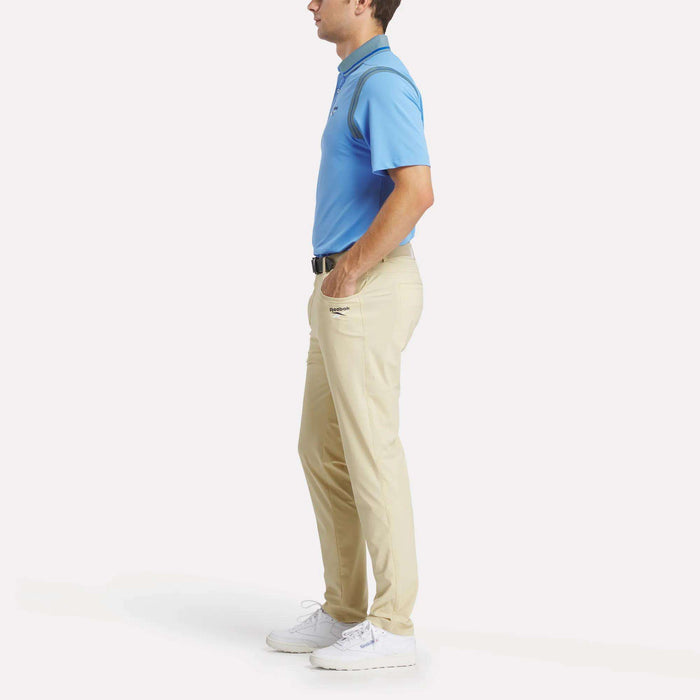 A man in profile wears a Reebok Men's Varsity Polo Shirt in light blue, beige pants, a black belt, and white sneakers, with hands in pockets against a plain white background.
