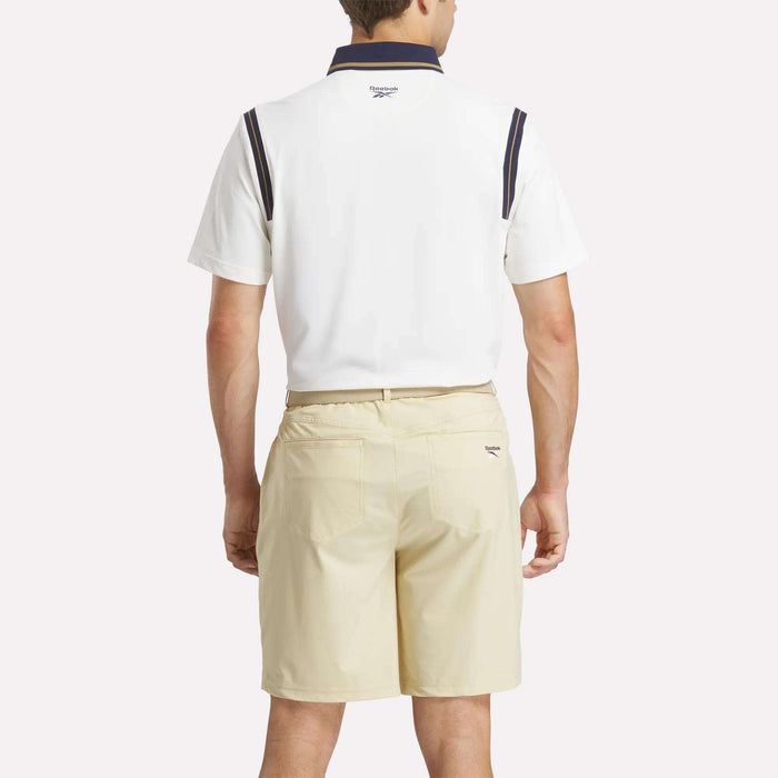 A man faces away, wearing a Reebok Men's Varsity Polo Shirt with dark trim on the collar and sleeves, paired with light beige shorts. The shirt is made with RBK-DRY fabric. He stands against a plain light background.