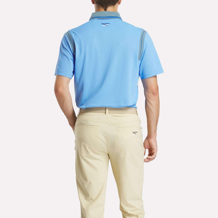 A man stands with his back to the camera, wearing a light blue Reebok Men's Varsity Polo Shirt and beige pants against a plain white background.