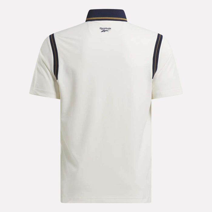Back view of the Men's Varsity Polo Shirt by Reebok, crafted with RBK-DRY fabric. Features a white short-sleeve design, navy collar, navy shoulder stripes, and a Reebok logo at the upper back below the collar.