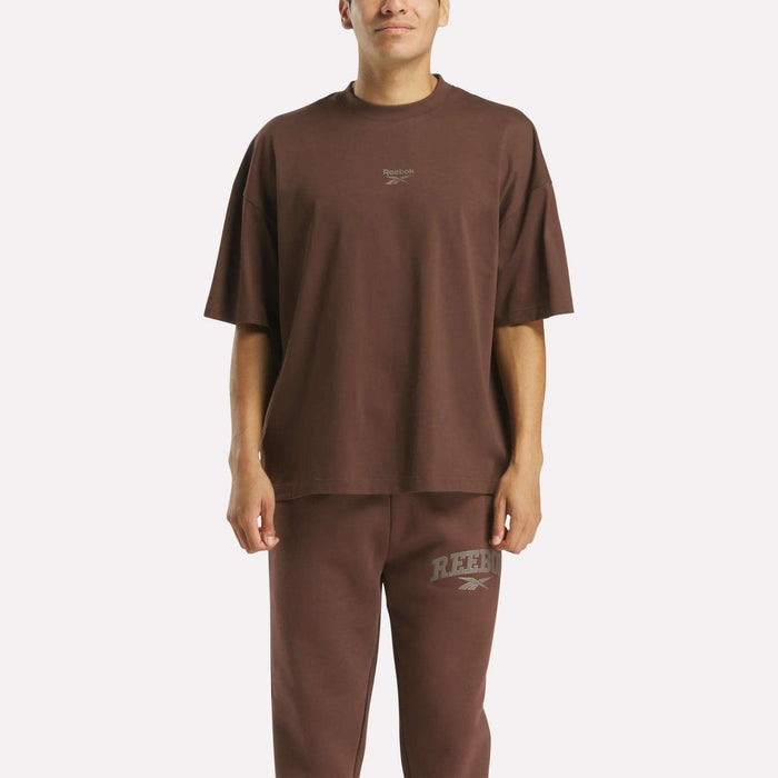 A person models an athletic look in the Men's Varsity T-Shirt and matching sweatpants by Reebok, both in loose-fitting dark brown with subtle brand logos, set against a plain white background.