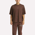 A person models an athletic look in the Men's Varsity T-Shirt and matching sweatpants by Reebok, both in loose-fitting dark brown with subtle brand logos, set against a plain white background.