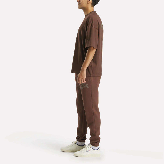 A person is shown in profile wearing a loose-fitting Reebok Men's Varsity T-Shirt paired with jogger pants and white sneakers, highlighting an athletic look against a plain white background.