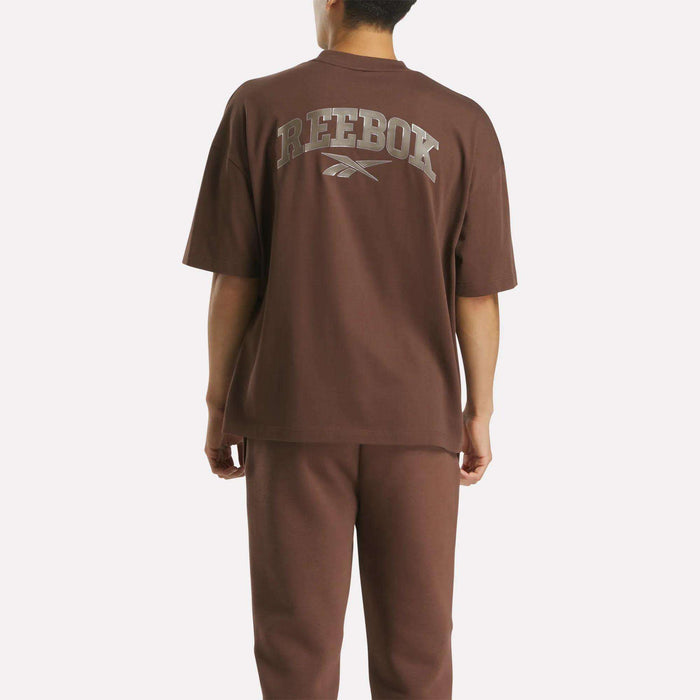 A person faces away, wearing the Reebok Men's Varsity T-Shirt and matching pants. The shirt displays REEBOK and the logo boldly across the back, giving an athletic edge to the outfit.