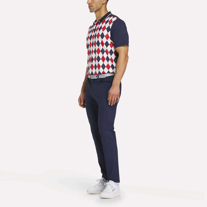 A man in a side pose wears the Reebok Men's Vector Argyle Print Polo with a red, white, and blue argyle pattern, paired with navy pants, a white belt, and white athletic shoes against a white background.