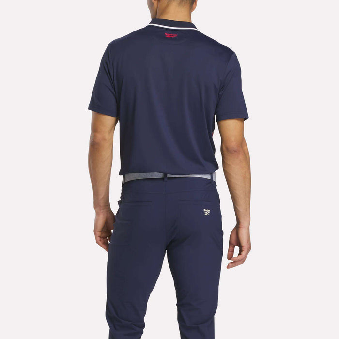 A man stands with his back to the camera, wearing a Reebok Men's Vector Argyle Print Polo in navy and matching navy pants with a light gray belt, both crafted from stretch knit fabric for superior comfort.