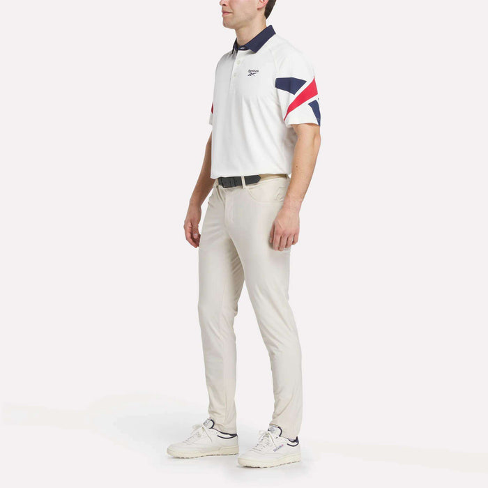 A man stands against a plain white background wearing a men's Reebok Vector Sleeve Polo Shirt with navy and red sleeve stripes, light-colored pants, a black belt, and white sneakers.