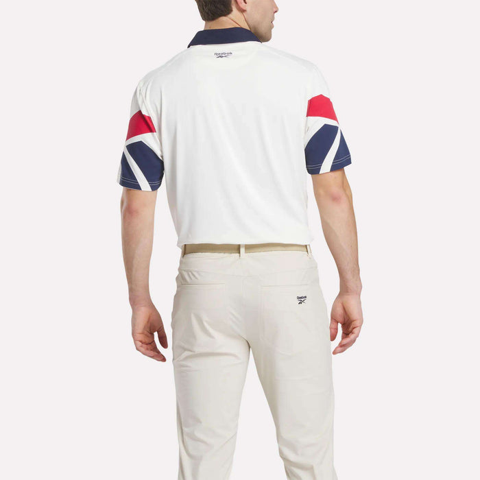 A man stands facing away, wearing the Reebok Men's Vector Sleeve Polo Shirt with UPF-rated stretch fabric, a white body, dark collar, red and blue geometric sleeve patterns, cream pants, all set against a plain white background.