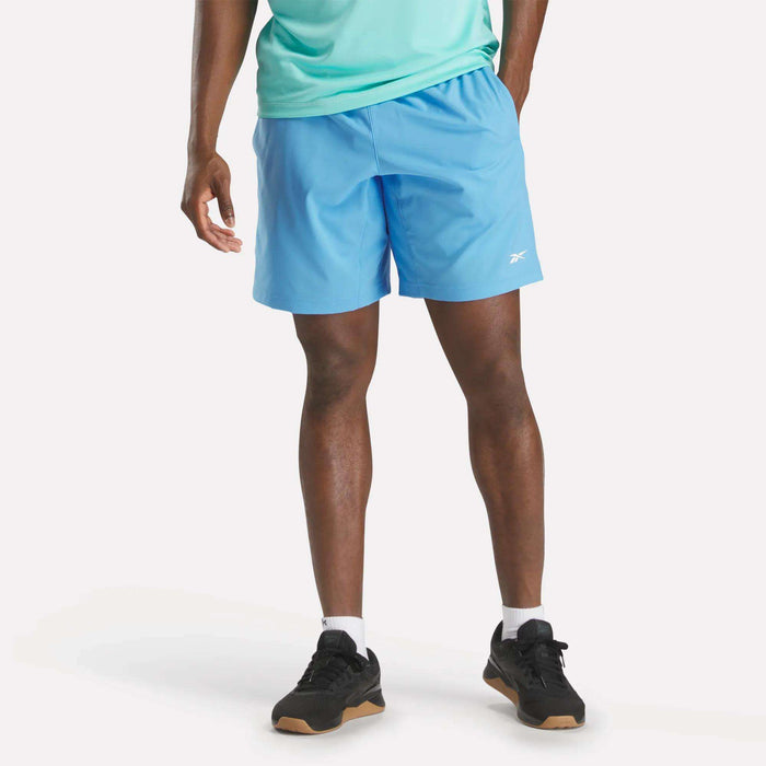A man wears a light blue T-shirt, Reebok Men's Workout Ready Shorts 9" in matching blue, black sneakers, and white socks, standing against a plain white background.