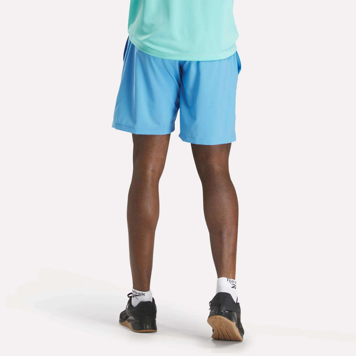 A person in a light teal shirt, Men's Workout Ready Shorts 9" by Reebok in blue, white socks, and black sneakers stands facing away, showing the back of their legs against a plain white background.