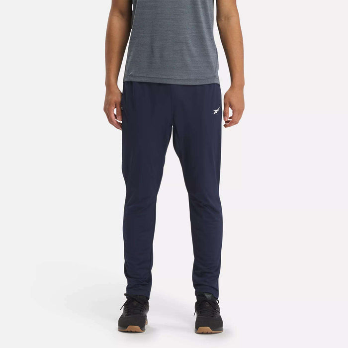 A person stands against a white background wearing Reebok Men's Workout Ready Track Pant in navy blue with a small white logo on the left thigh, paired with a grey short-sleeve shirt and black sneakers.