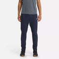 A person stands against a white background wearing Reebok Men's Workout Ready Track Pant in navy blue with a small white logo on the left thigh, paired with a grey short-sleeve shirt and black sneakers.