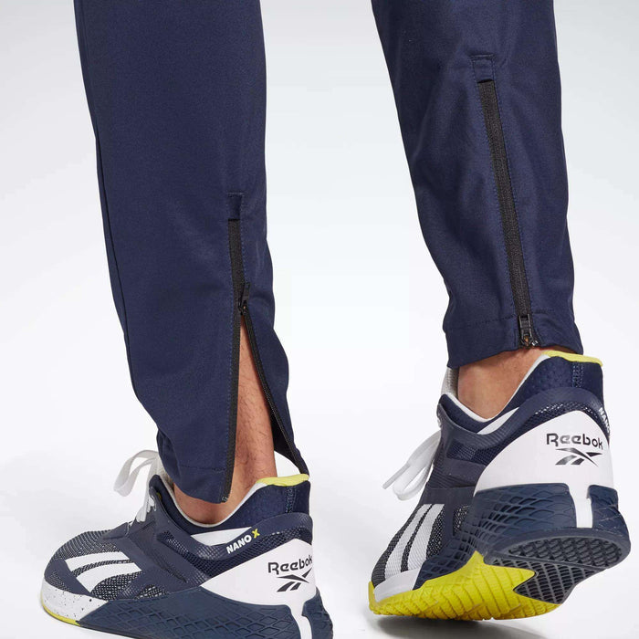 Close-up of a person in Reebok Men's Workout Ready Track Pant with ankle zippers and navy Reebok sneakers featuring white and yellow soles, focusing on the back of the shoes and lower legs.