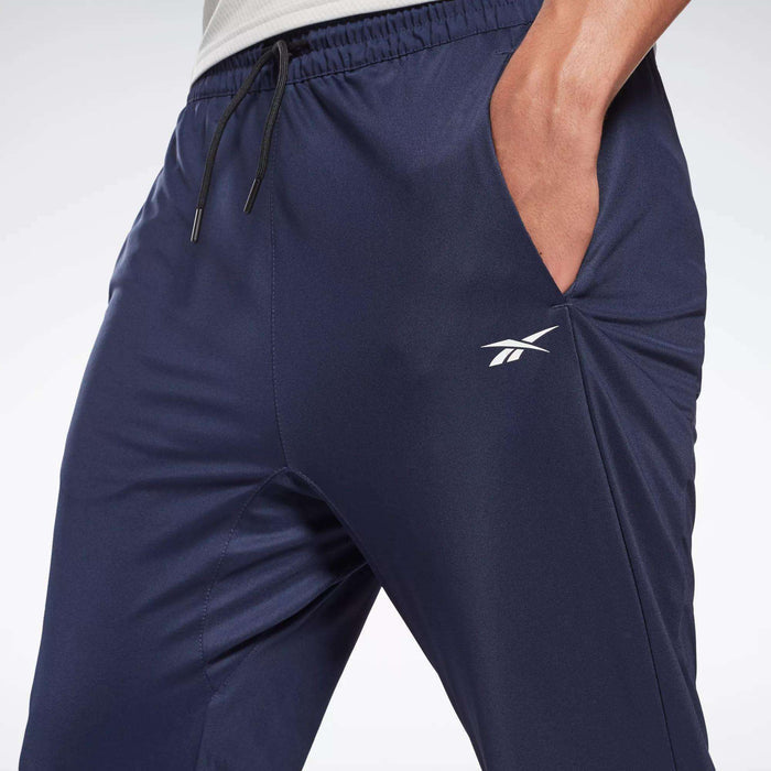 Close-up of a person wearing Reebok Men's Workout Ready Track Pant in navy blue, featuring the white Reebok logo on the left thigh and their right hand in the pocket.