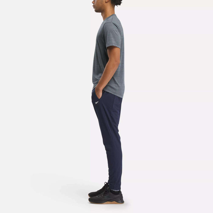 A man in profile stands with his hands in his pockets, wearing a gray t-shirt, Reebok Men's Workout Ready Track Pant in navy, and black athletic shoes against a plain, light background.