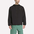 A man wears a black Reebok Men's Woven Jacket with a relaxed fit and long sleeves, paired with light green shorts, standing against a plain white background.