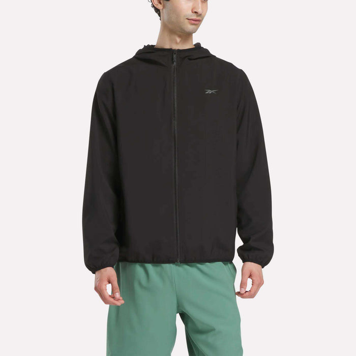 A man wears a black Reebok Men's Woven Jacket with a relaxed fit and long sleeves, paired with light green shorts, standing against a plain white background.