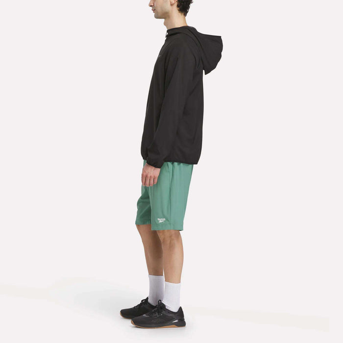 A person standing sideways in a Reebok Men's Woven Jacket, green shorts, white crew socks, and black athletic shoes against a plain white background.