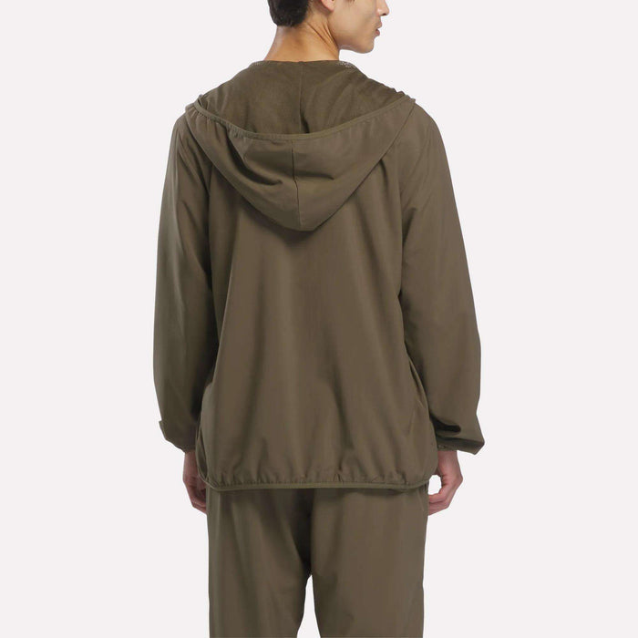 A person stands with their back to the camera, wearing the Reebok Men’s Woven Jacket in olive green along with matching pants. The lightweight jacket features a loose fit and the hood rests down, set against a plain, light-colored background that highlights the streamlined aesthetic of men’s training apparel.