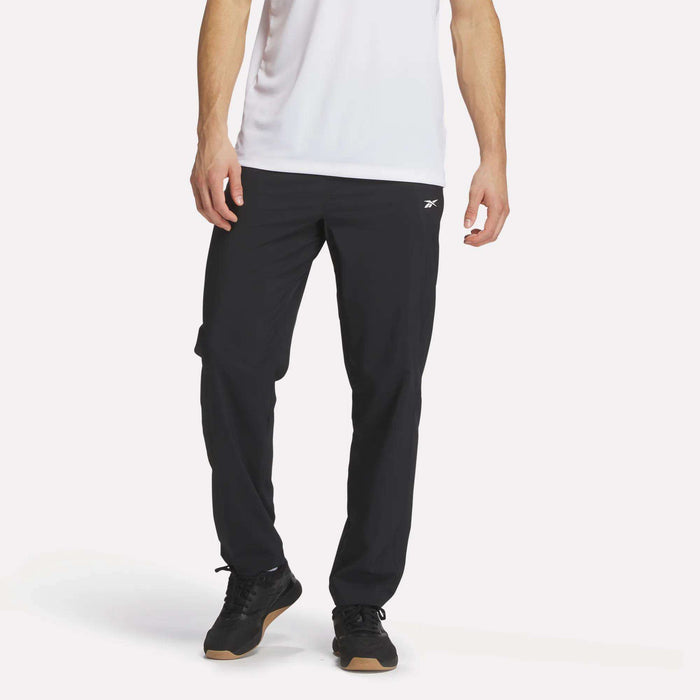 A person wears a white short-sleeve shirt and Reebok Men's Woven Pants, featuring a small white logo, with black sneakers, standing against a plain white background.