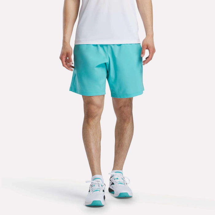 A person, head not visible, stands against a plain white background wearing a white shirt, Reebok Men's Woven Shorts in turquoise, and white sneakers with turquoise accents.