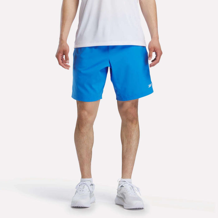 A person wearing a white shirt, Reebok Men's Woven Shorts in bright blue, and white sneakers stands facing forward against a plain white background. The image is cropped at the shoulders.