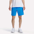 A person wearing a white shirt, Reebok Men's Woven Shorts in bright blue, and white sneakers stands facing forward against a plain white background. The image is cropped at the shoulders.