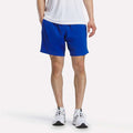 A person wearing a white shirt, Reebok Men's Woven Shorts in bright blue, and white sneakers stands against a plain background. Only the lower body is visible.