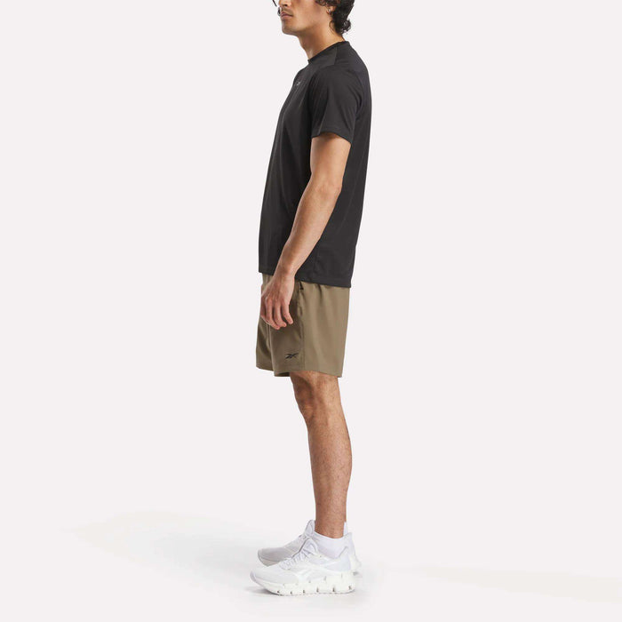 A man stands in profile against a plain background, wearing a black T-shirt, Reebok Men's Workout Ready Woven Shorts in khaki, and white athletic shoes.