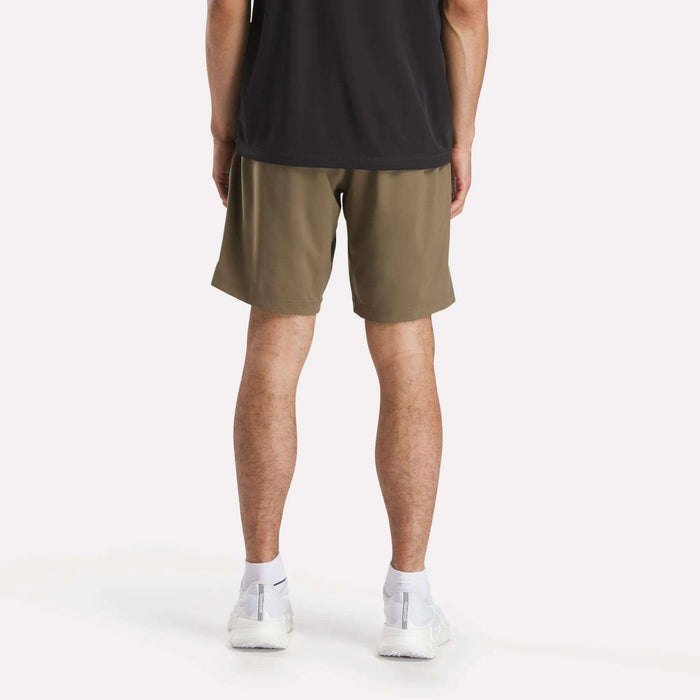 A person stands facing away against a plain light background, wearing a black shirt, white athletic shoes, and olive green Reebok Men's Workout Ready Woven Shorts.