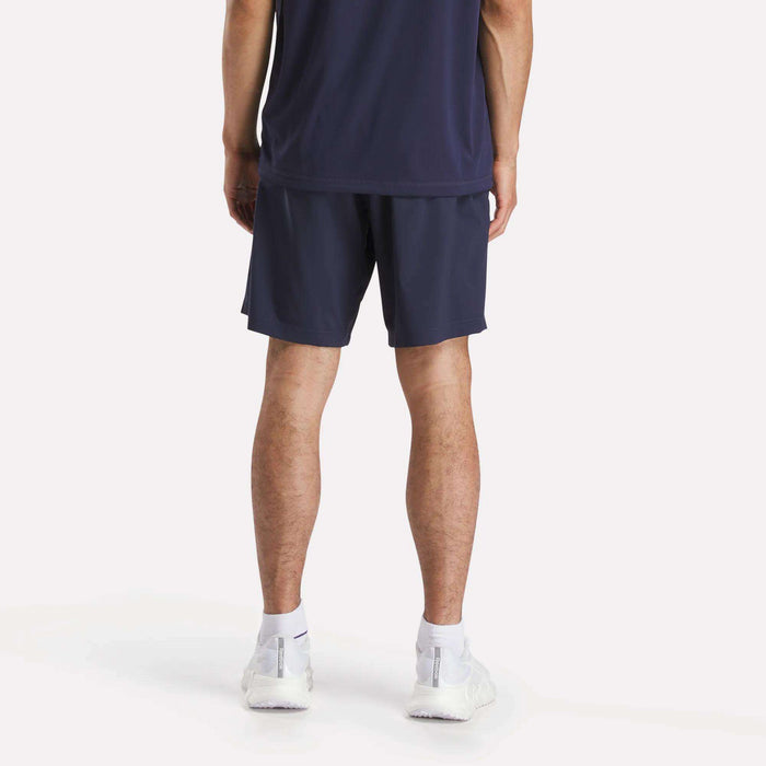 A person stands facing away from the camera, wearing a navy athletic shirt and Reebok Men's Workout Ready Woven Shorts, paired with white socks and sneakers against a plain white background.