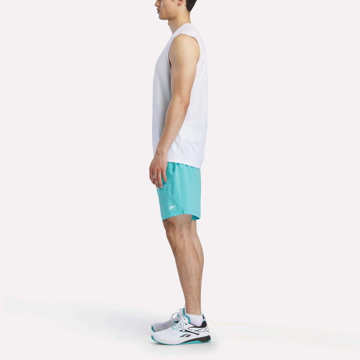 A man stands in profile wearing a white sleeveless shirt, Men’s Woven Shorts by Reebok in light blue, and white sneakers with black and teal accents, set against a plain white background.