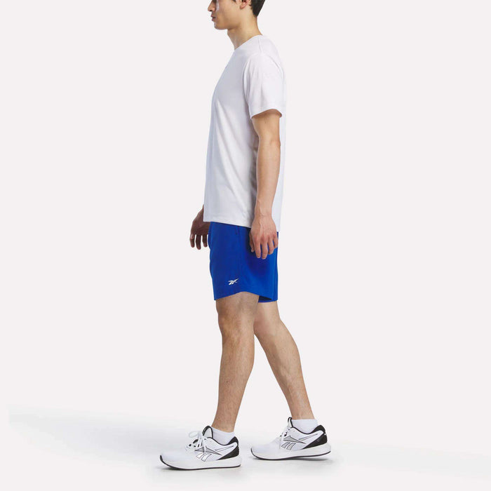 A man in profile wears a white T-shirt, Reebok Men's Woven Shorts in bright blue, and white sneakers with black details, set against a plain white background.