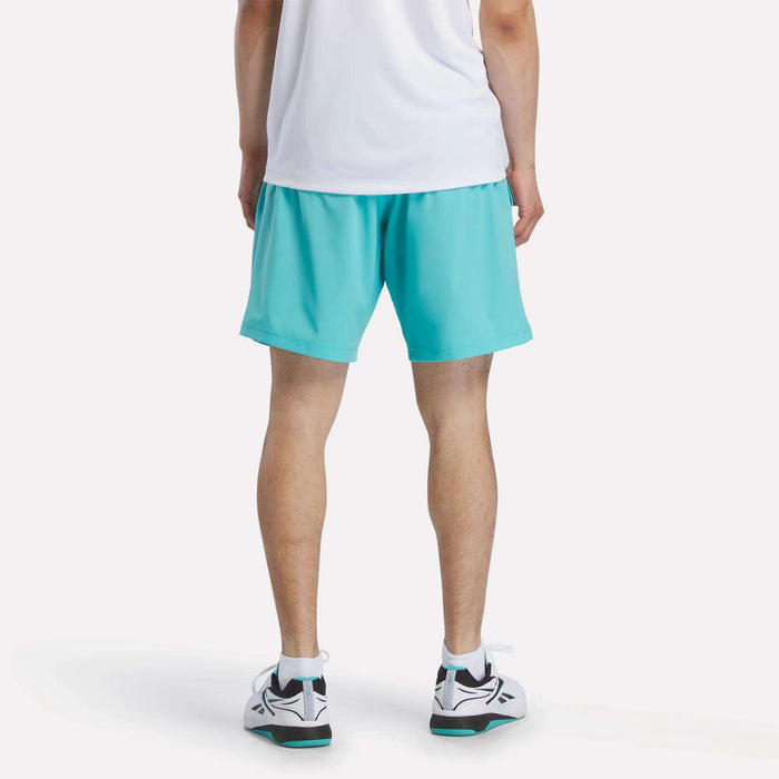 A person faces away in a white shirt, Reebok Men's Woven Shorts in turquoise, and white sneakers with black and teal accents. The plain, light-colored background keeps the focus on the outfit.